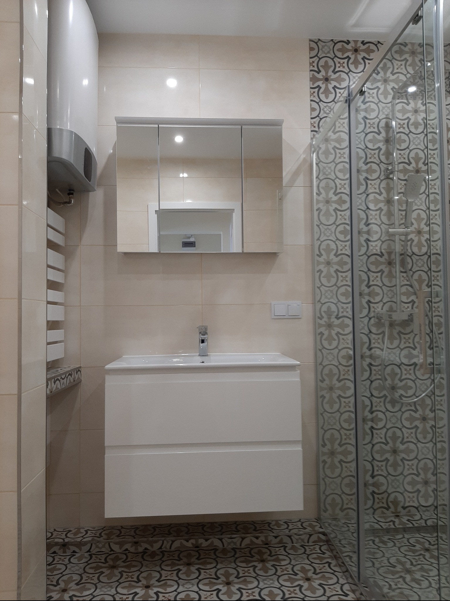 Wykończona łazienka z kabiną prysznicową, umywalką z szafką i grzejnikiem. Beżowe płytki na ścianach i wzorzysta podłoga. Lustro z szafką nad umywalką.