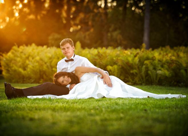 Para młoda leżąca na trawie w promieniach zachodzącego słońca, panna młoda w sukni z długim trenem opiera głowę na kolanach pana młodego ubranego w białą koszulę i muszkę.