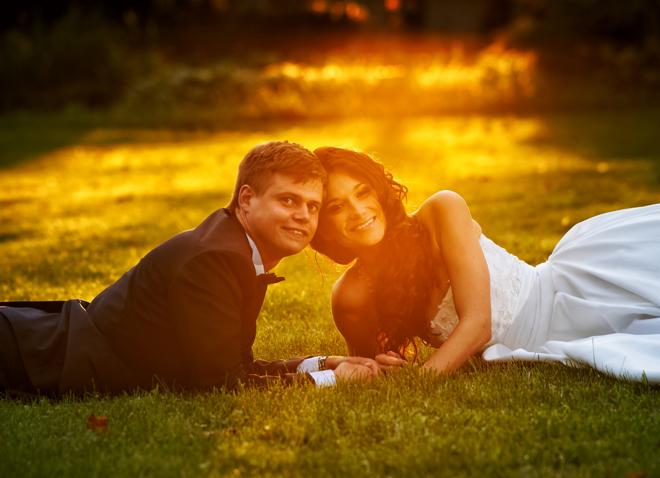 Uśmiechnięta para młoda, leżąca na trawie w promieniach zachodzącego słońca, trzymająca się za ręce.