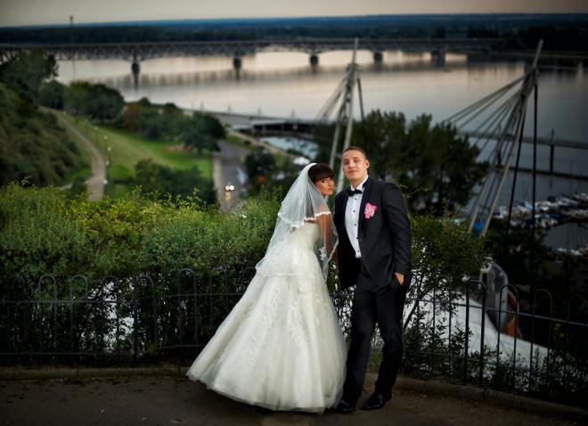 Para młoda pozuje na tle panoramy miasta z rzeką i mostami, panna młoda w białej sukni z welonem, pan młody w czarnym garniturze z różową butonierką.