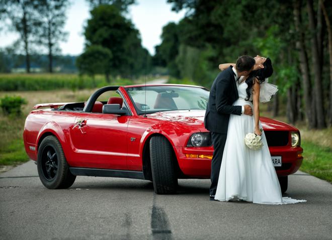 Para młoda w objęciach na tle czerwonego kabrioletu Ford Mustang, bukiet ślubny w dłoni panny młodej, wiejska droga w tle.