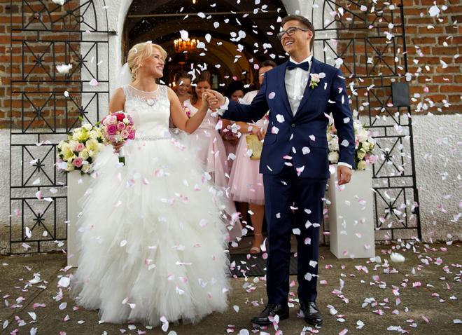 Uśmiechnięta para młoda wychodzi z kościoła podczas deszczu płatków róż, panna młoda w białej sukni z bukietem, pan młody w granatowym garniturze i okularach, w tle druhny i dekoracje kwiatowe.
