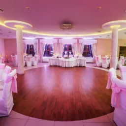 Elegancko udekorowana sala balowa z drewnianym parkietem, białymi krzesłami z różowymi kokardami, białymi obrusami i oświetleniem w ciepłych barwach.