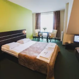 Pokój hotelowy z dwoma łóżkami, stolikiem z krzesłami i telewizorem, utrzymany w stonowanej kolorystyce z akcentem zielonej ściany.