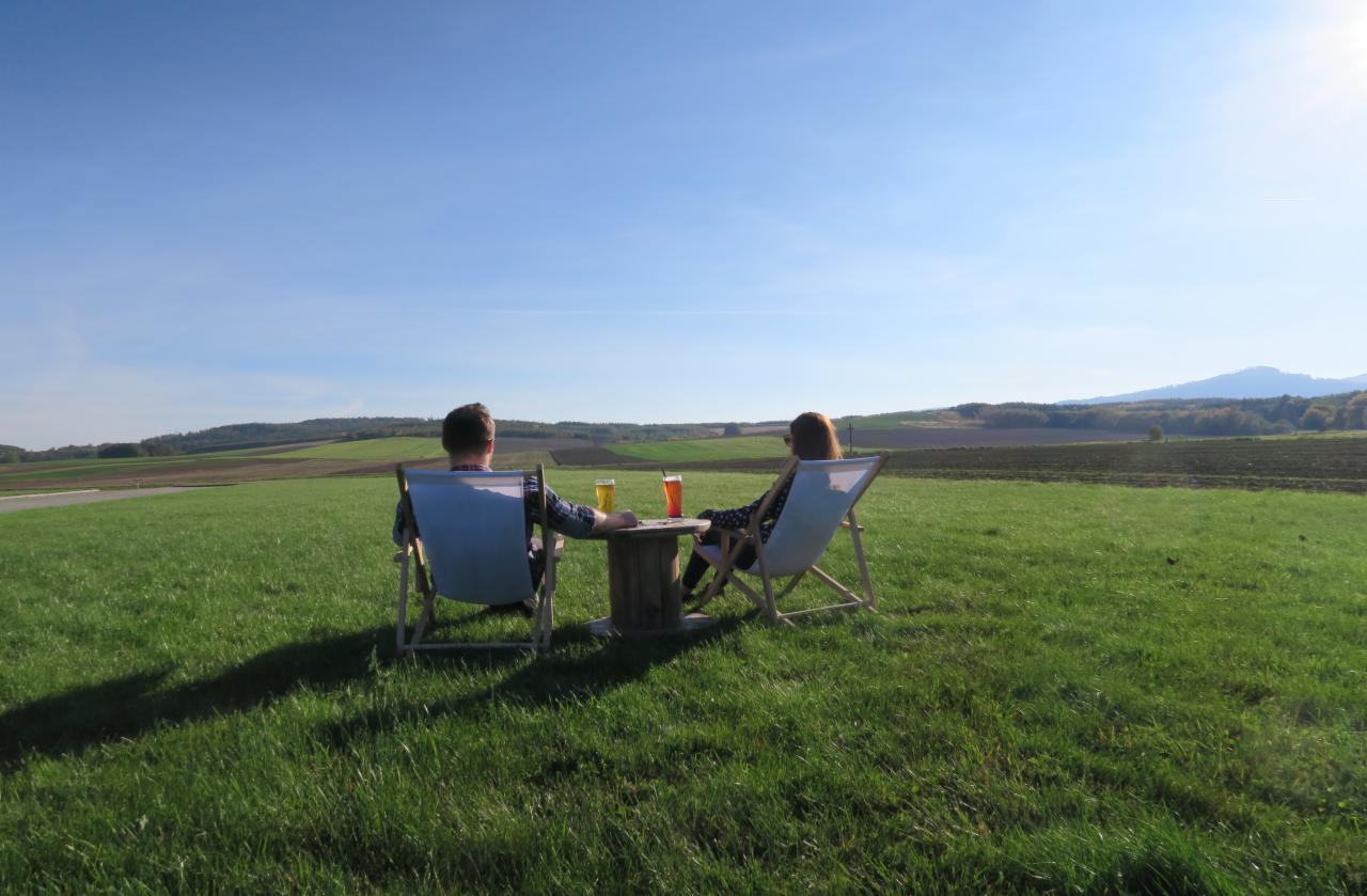 Para relaksuje się w leżakach na trawie, z napojami na drewnianym stoliku, widok na pola i wzgórza w słoneczny dzień.