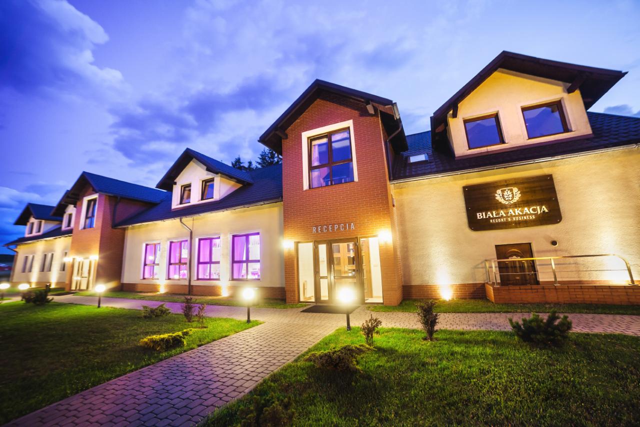 Elewacja budynku 'Biała Akacja Resort & Business' o zmierzchu, z oświetlonym wejściem do recepcji i brukowanym chodnikiem prowadzącym przez trawnik.
