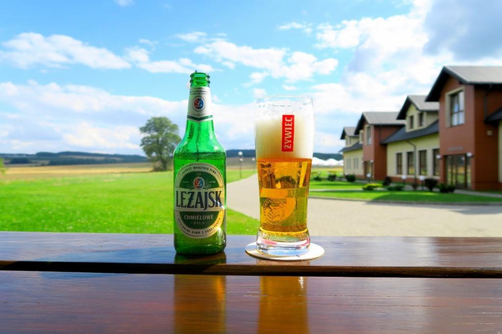 Butelka piwa Leżajsk i szklanka piwa Żywiec na drewnianym stole z widokiem na zielone pola i zabudowania w tle, błękitne niebo z chmurami.