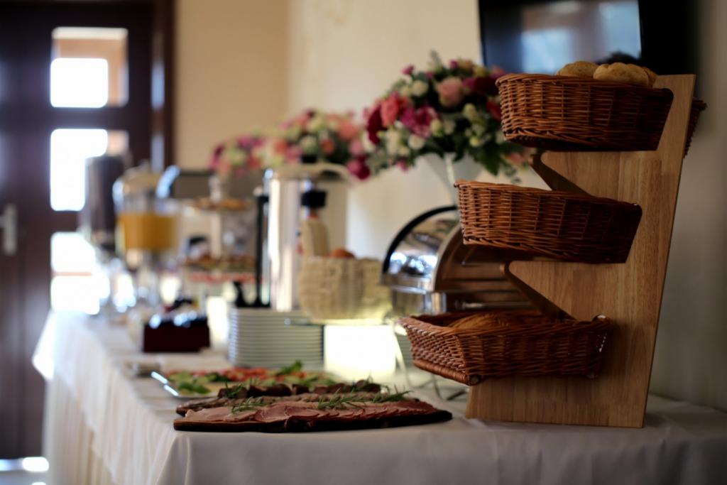 Stół bufetowy z przekąskami, wędlinami, sałatkami i pieczywem w wiklinowych koszykach na drewnianym stojaku, kwiaty w tle.