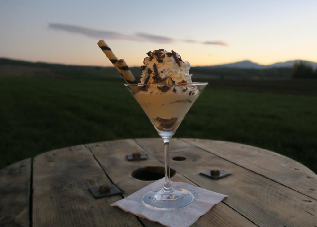 Deser tiramisu w szklance koktajlowej, udekorowany bitą śmietaną, posypką z czekolady i waflem, ustawiony na drewnianym stole z zielonym tłem krajobrazu o zmierzchu.