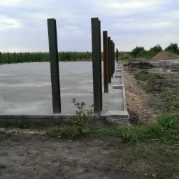 Wylana betonowa płyta fundamentowa z wystającymi stalowymi słupami, widok na pole kukurydzy i zarośla w tle, teren budowy.