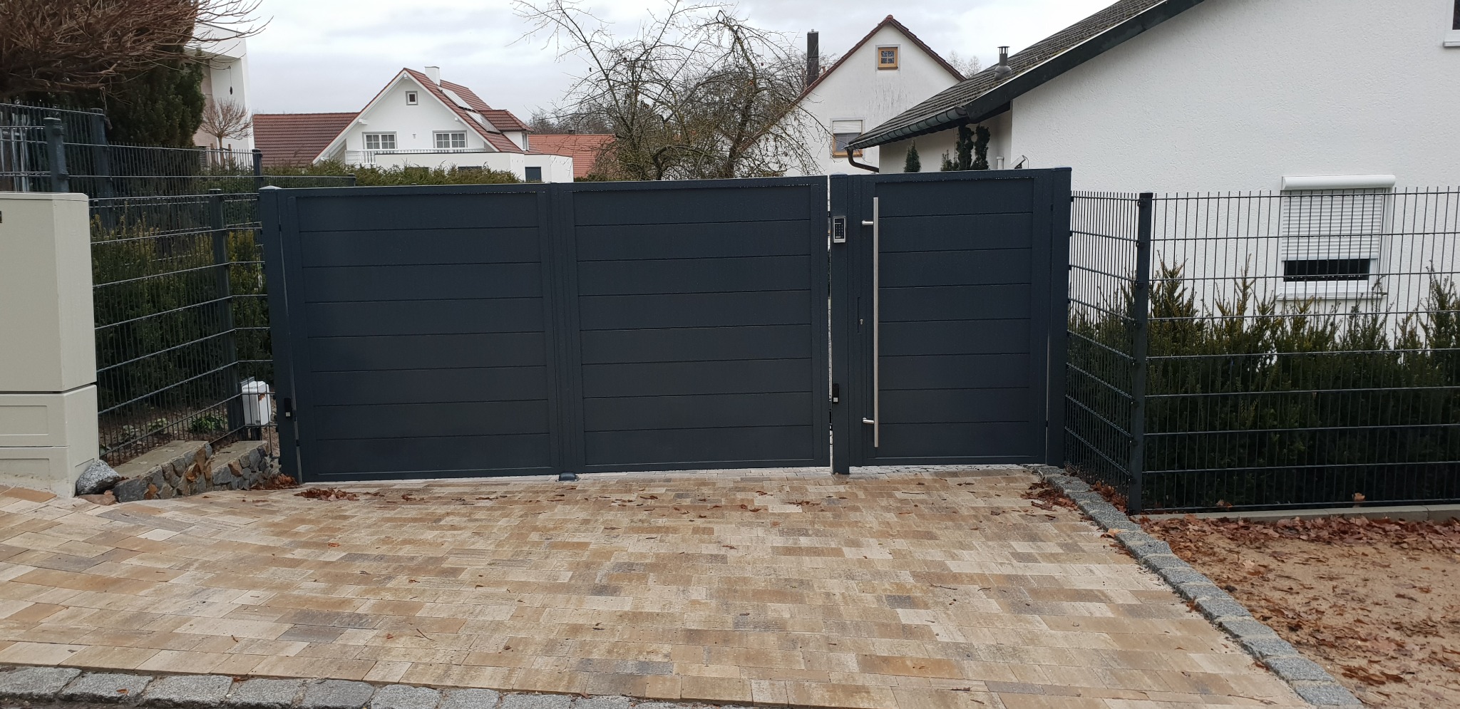Nowoczesna, pozioma, antracytowa brama wjazdowa z panelem sterowania i długim uchwytem ze stali nierdzewnej, sąsiadująca z metalowym ogrodzeniem panelowym i brukowanym podjazdem z kostki...