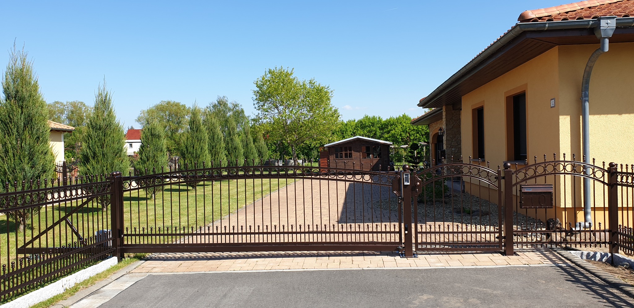 Brązowa, metalowa brama wjazdowa z automatyką i domofonem na tle zadbanego ogrodu i domu z pomarańczową elewacją.