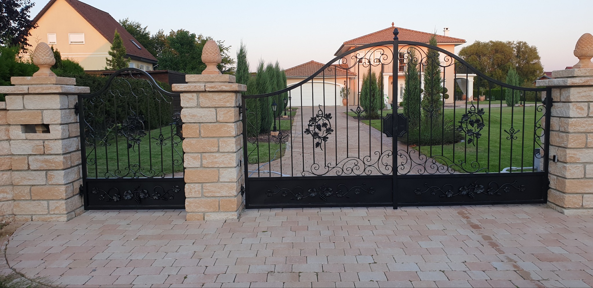 Czarna, ozdobna brama wjazdowa z motywem winogron, osadzona na słupkach z jasnego kamienia zwieńczonych dekoracyjnymi szyszkami, prowadząca do domu z ogrodem.