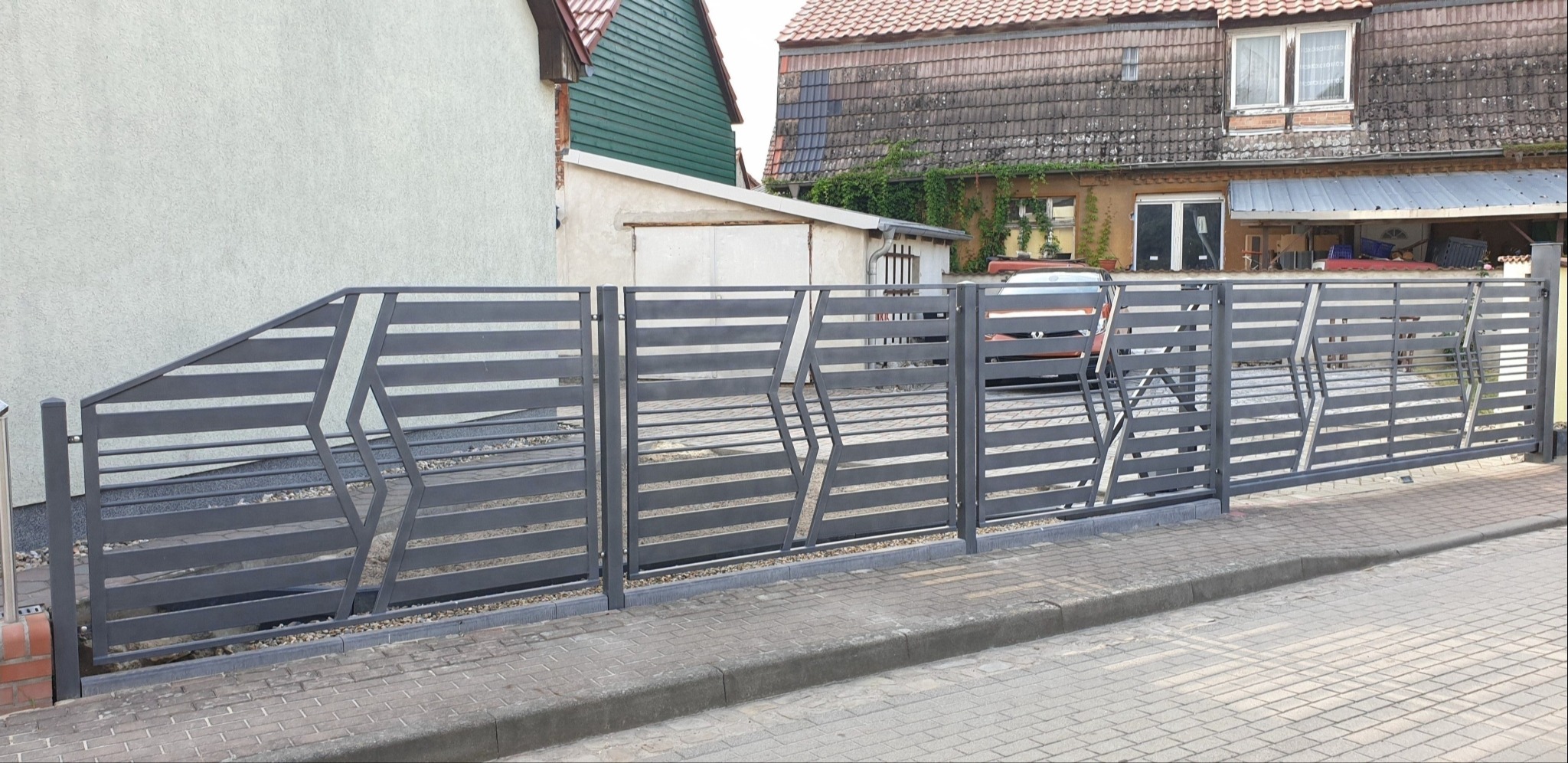 Nowoczesna, przesuwna brama wjazdowa z poziomymi panelami i geometrycznym wzorem w kształcie rombów, na tle zabudowań mieszkalnych.