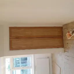 Dwa poziome, drewniane panele zabudowane nad oknem w trakcie remontu pomieszczenia; widoczne uszkodzone deski podłogowe i grzejnik.