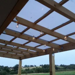 Drewniana pergola z przezroczystym zadaszeniem i dekoracyjnymi żarówkami w stylu retro, widok od dołu na tle zieleni.