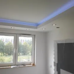 Białe wnętrze z oknem i podwieszanym sufitem z niebieskim oświetleniem LED, widoczne reflektory w suficie, drzwi zabezpieczone folią ochronną.