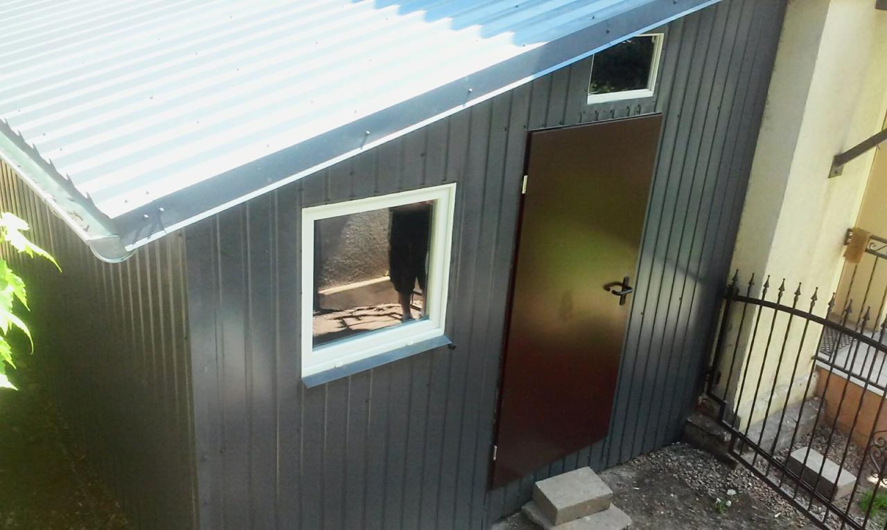 Metalowy budynek gospodarczy z falistej blachy, szarych ścianach i metalowym dachem, brązowe drzwi i białe okno, widok z góry.