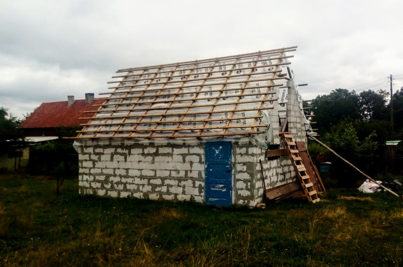 Budynek z pustaków z niebieskimi drzwiami w trakcie budowy dachu, widoczne łaty, membrana dachowa i drabina oparta o ścianę szczytową, w tle dom z czerwoną dachówką.
