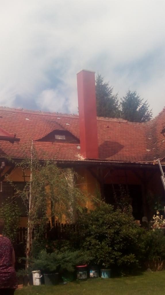 Czerwony komin po malowaniu na tle dachu z dachówki ceramicznej, widoczne ślady prac na dachu i farba w wiadrach na trawniku.