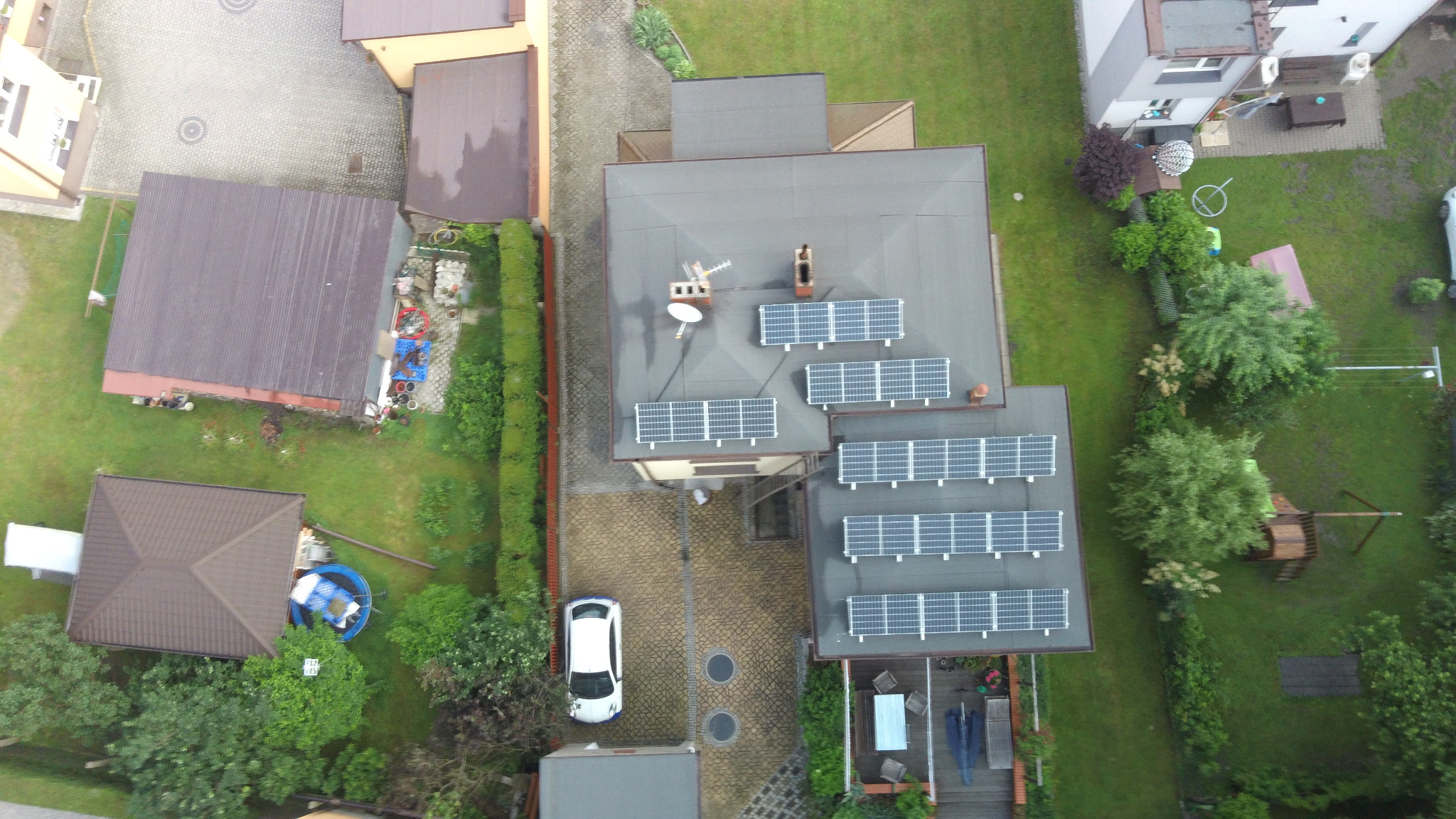 Widok z lotu ptaka na dom z panelami słonecznymi na dachu, antena satelitarna, biały samochód na podjeździe z kostki brukowej, zielony ogród z trampoliną i placem zabaw