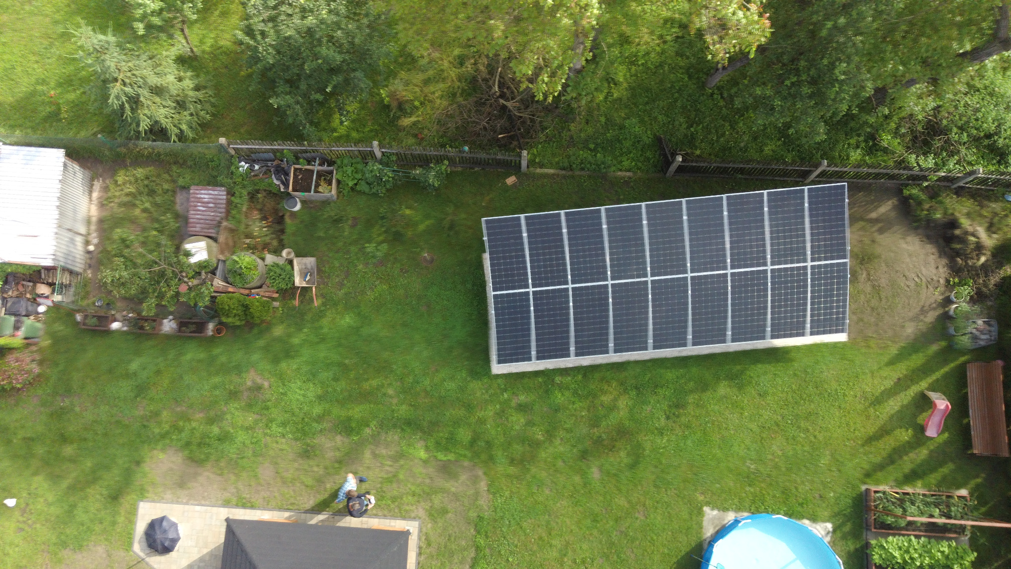 Widok z góry na panele słoneczne w ogrodzie, z widocznym basenem i zaroślami w tle.