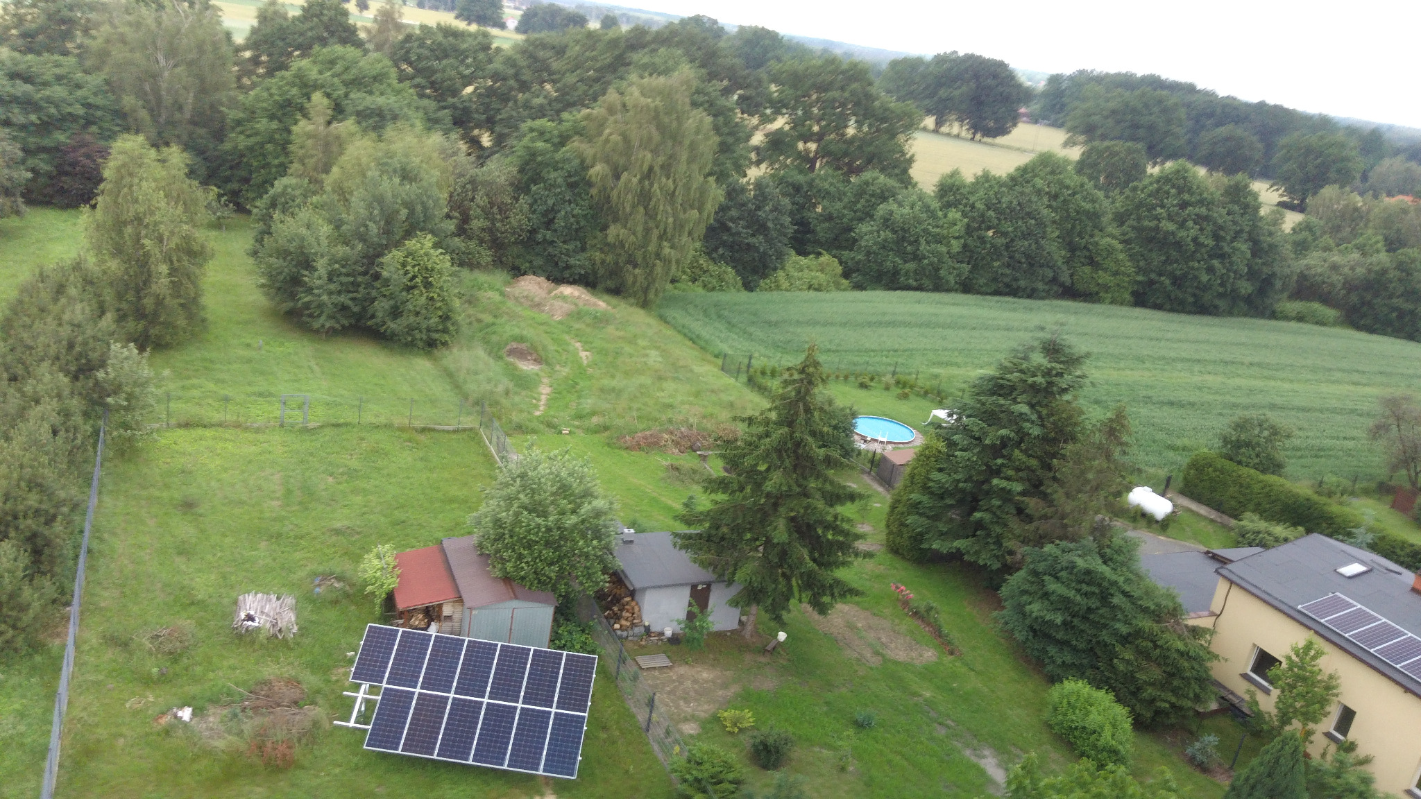 Widok z lotu ptaka na posesję z zainstalowanymi panelami fotowoltaicznymi na ziemi obok zabudowań gospodarczych, w tle dom z panelami na dachu oraz basen.