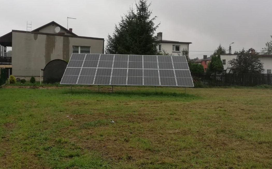 Panel fotowoltaiczny na trawniku przed domem w pochmurny dzień. Widoczne budynki w tle.