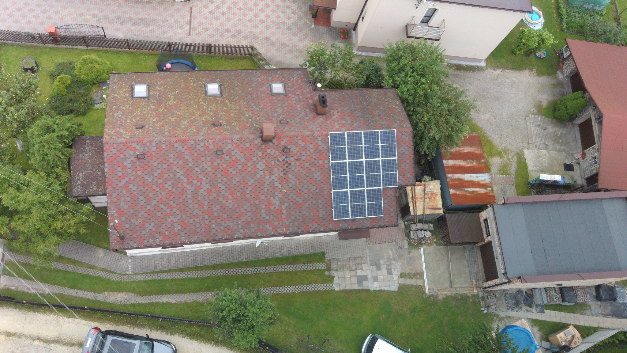 Widok z góry na dom z czerwoną dachówką i zamontowanymi panelami słonecznymi, otoczony zielenią i innymi budynkami.