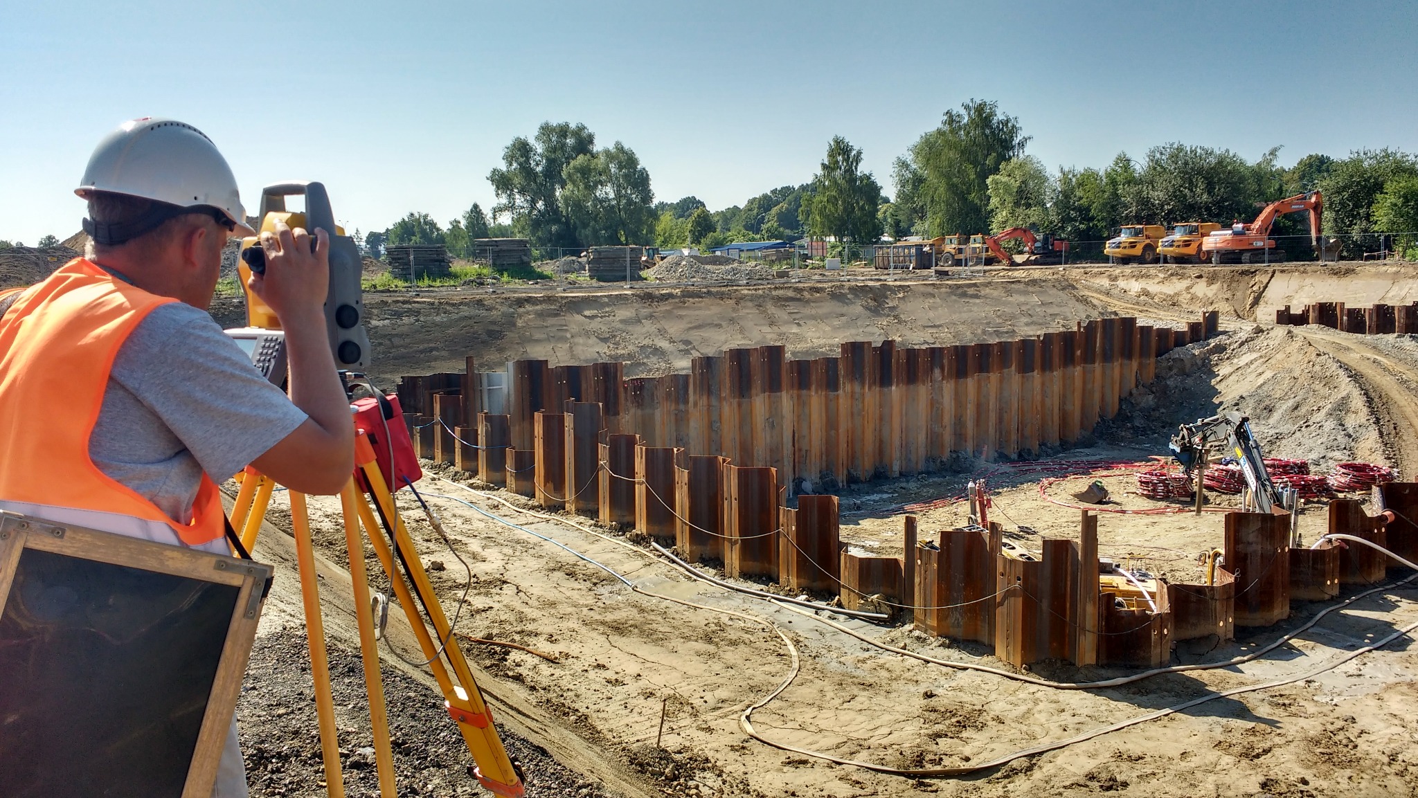 Praca geodezyjna na budowie: geodeta w kasku i kamizelce mierzy teren teodolitem, w tle wykop zabezpieczony ścianką Larsena i ciężki sprzęt.