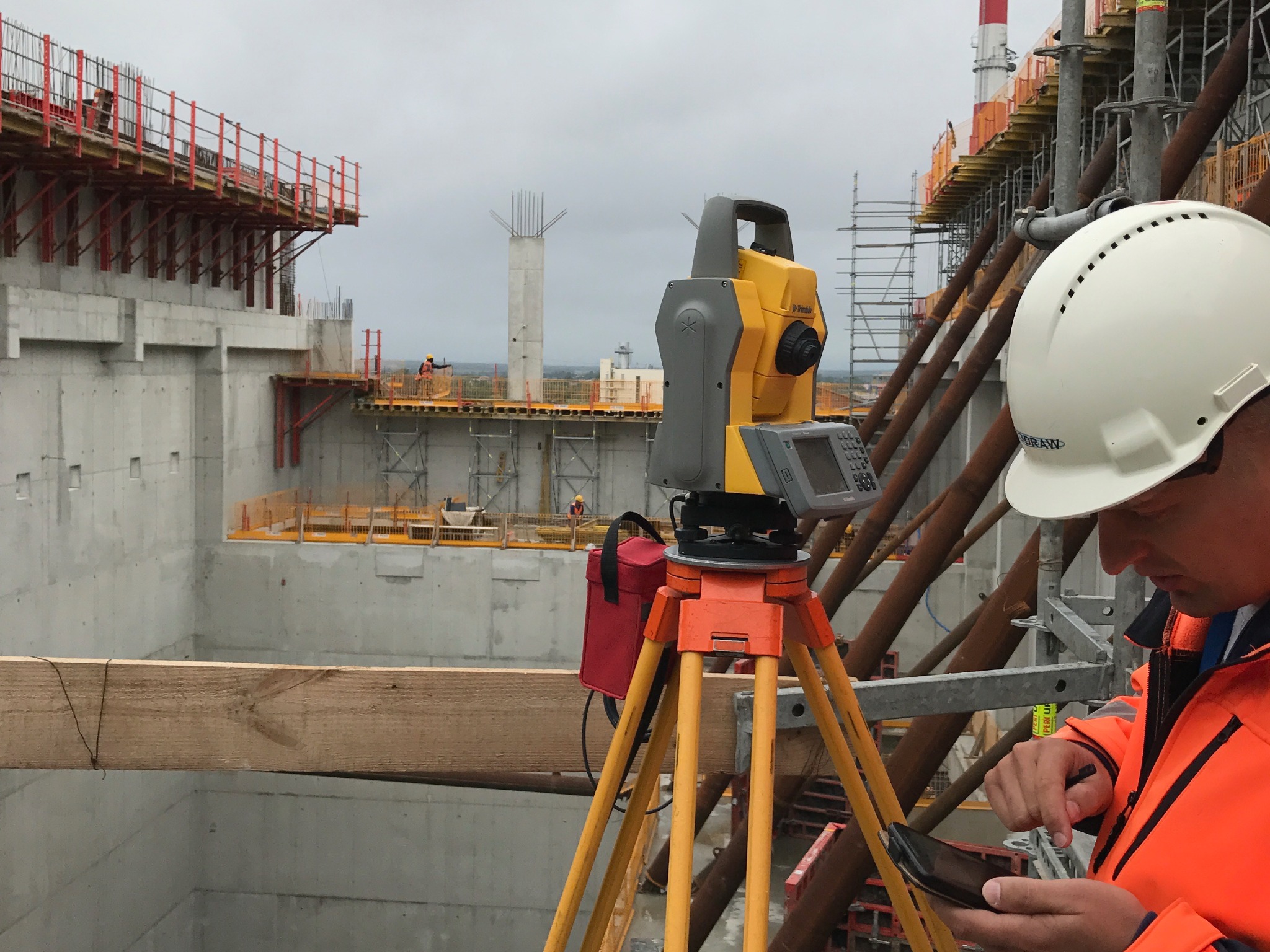 Praca geodezyjna na budowie: teodolit na statywie, pracownik w kasku i pomarańczowej kurtce obsługuje urządzenie mobilne. W tle surowy beton i elementy konstrukcyjne.