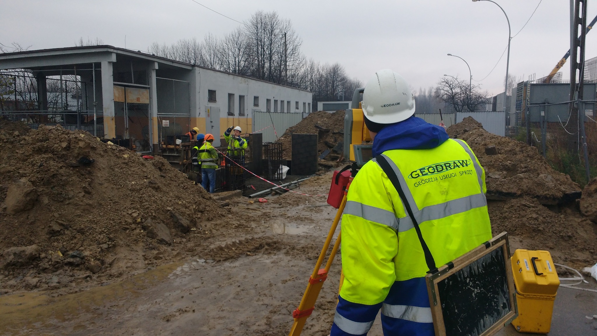 Praca geodezyjna na budowie: geodeta z niwelatorem na tle wykopu i grupy robotników w odblaskowych kamizelkach. Widoczny budynek w tle i błotniste podłoże.