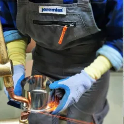 Spawanie punktowe metalowego elementu przez pracownika w rękawicach ochronnych, w tle zawieszone elementy oraz budynek firmy z logo Jeremias.