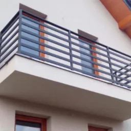 Nowoczesna, pozioma, metalowa barierka balkonowa w kolorze grafitowym, zamontowana na balkonie budynku mieszkalnego z widocznymi oknami.