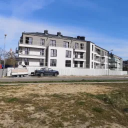 Nowoczesny budynek wielorodzinny z białymi elewacjami i szarymi elementami, częściowo ogrodzony białym płotem, z zaparkowanym pickupem z przyczepą i robotnikami w tle.