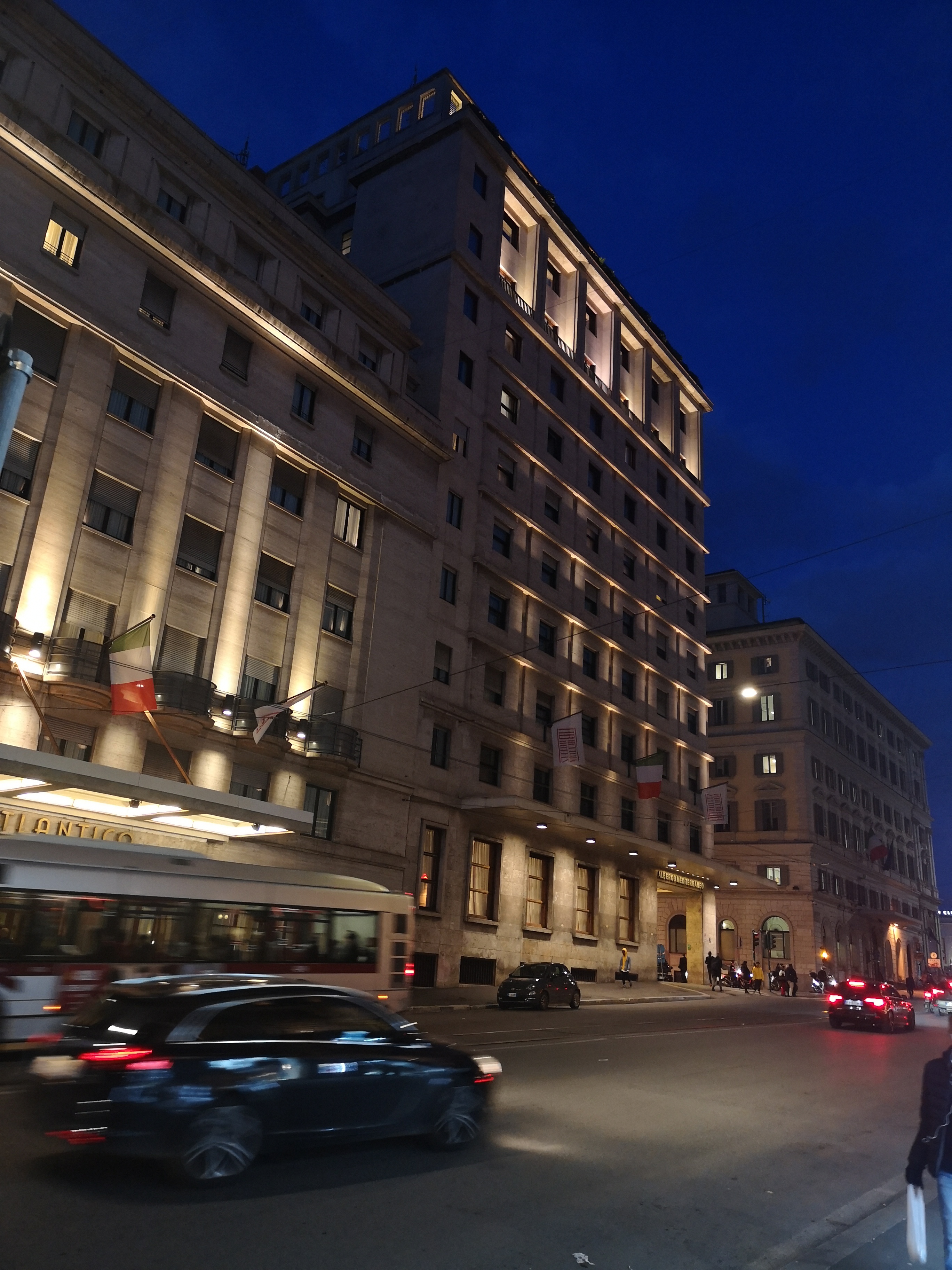 Elewacja kamienicy z oświetleniem nocnym, widoczne flagi i ruch uliczny w tle.