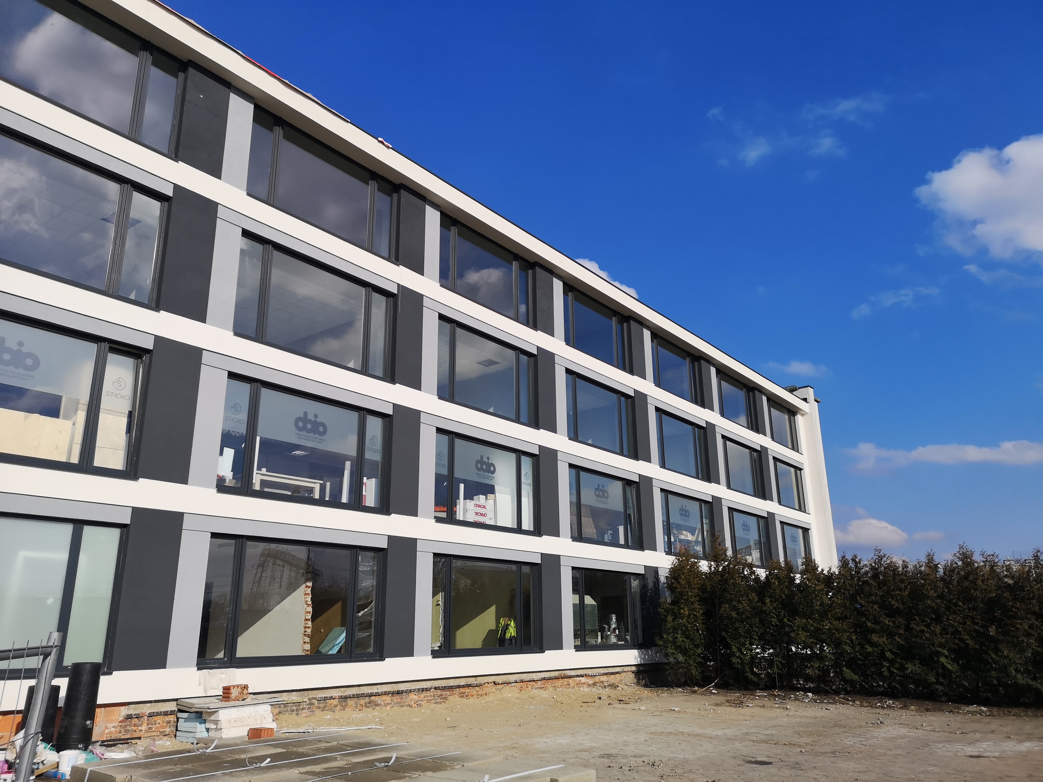 Nowoczesny budynek biurowy z elewacją w odcieniach szarości i bieli, z dużymi oknami z naklejonymi logo firm, widok z zewnątrz na plac budowy z ziemią, cegłami i materiałami budowlanymi, na tle...