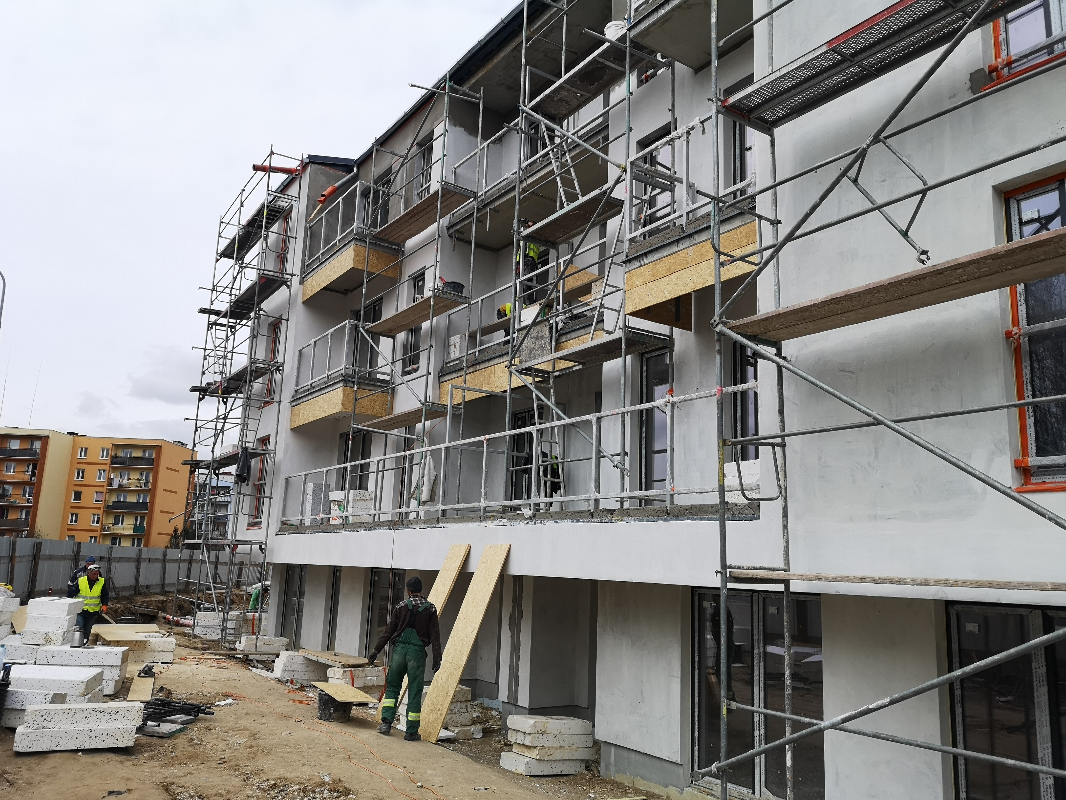 Budynek wielorodzinny w trakcie prac elewacyjnych, widoczne rusztowania, ocieplenie balkonów płytami OSB, pracownicy w odblaskowych kamizelkach i stosy bloczków betonowych na placu budowy.
