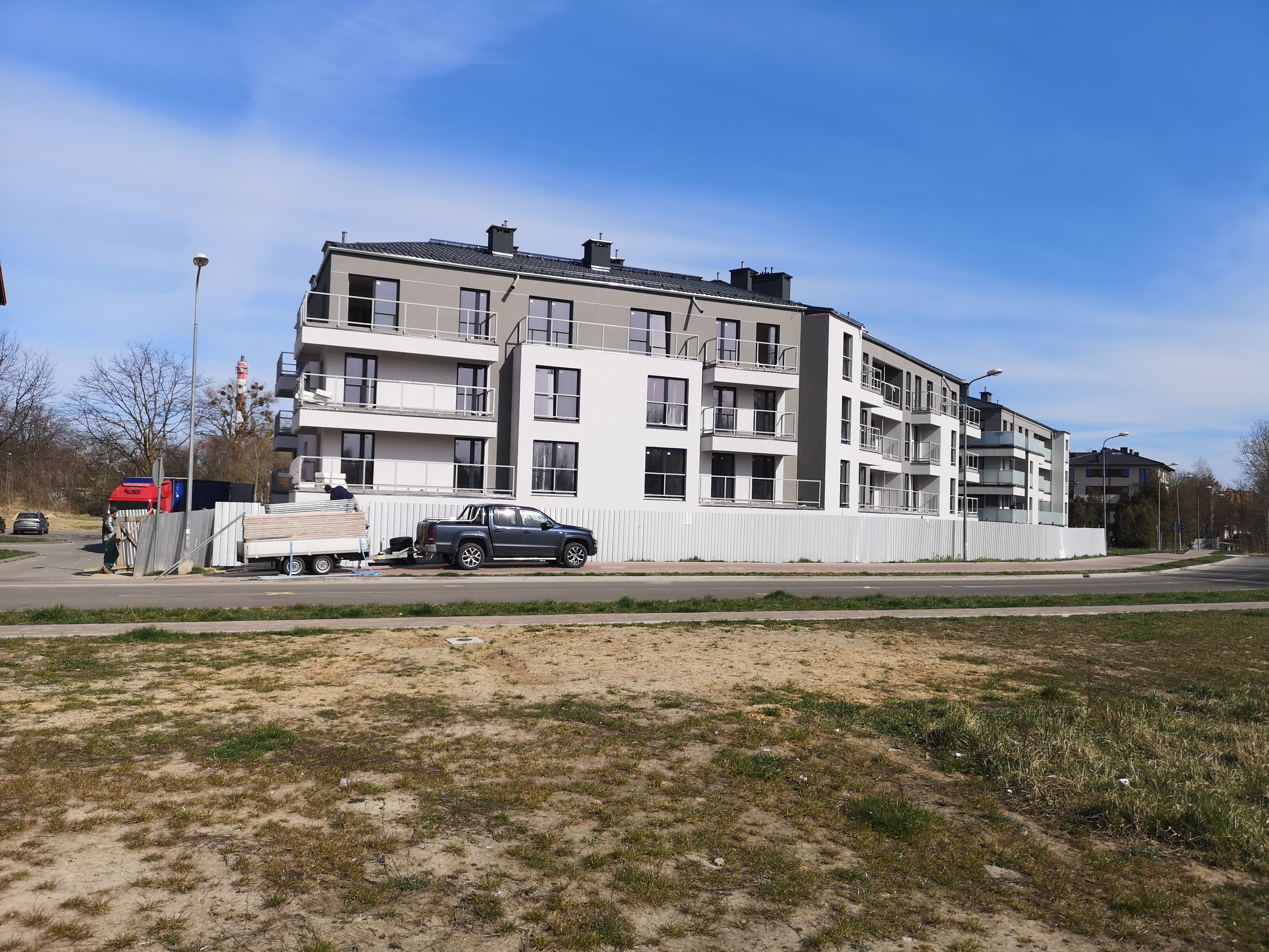 Nowoczesny budynek wielorodzinny z białymi elewacjami i szarymi elementami, częściowo ogrodzony białym płotem, z zaparkowanym pickupem z przyczepą i robotnikami w tle.