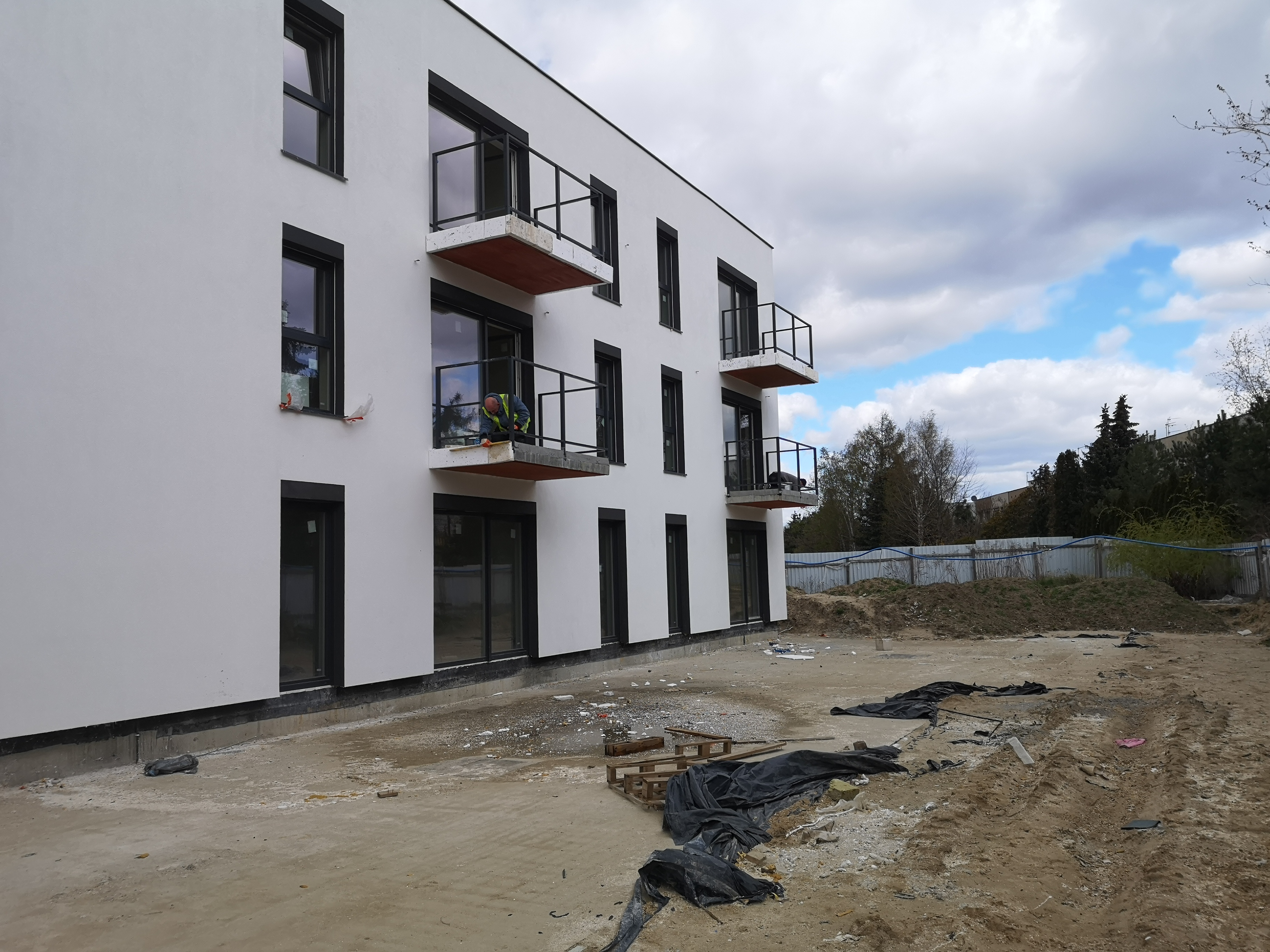 Pracownik w kamizelce na balkonie nowego budynku, montuje balustradę, widoczne okna z czarnymi ramami i biała elewacja, teren budowy w tle.