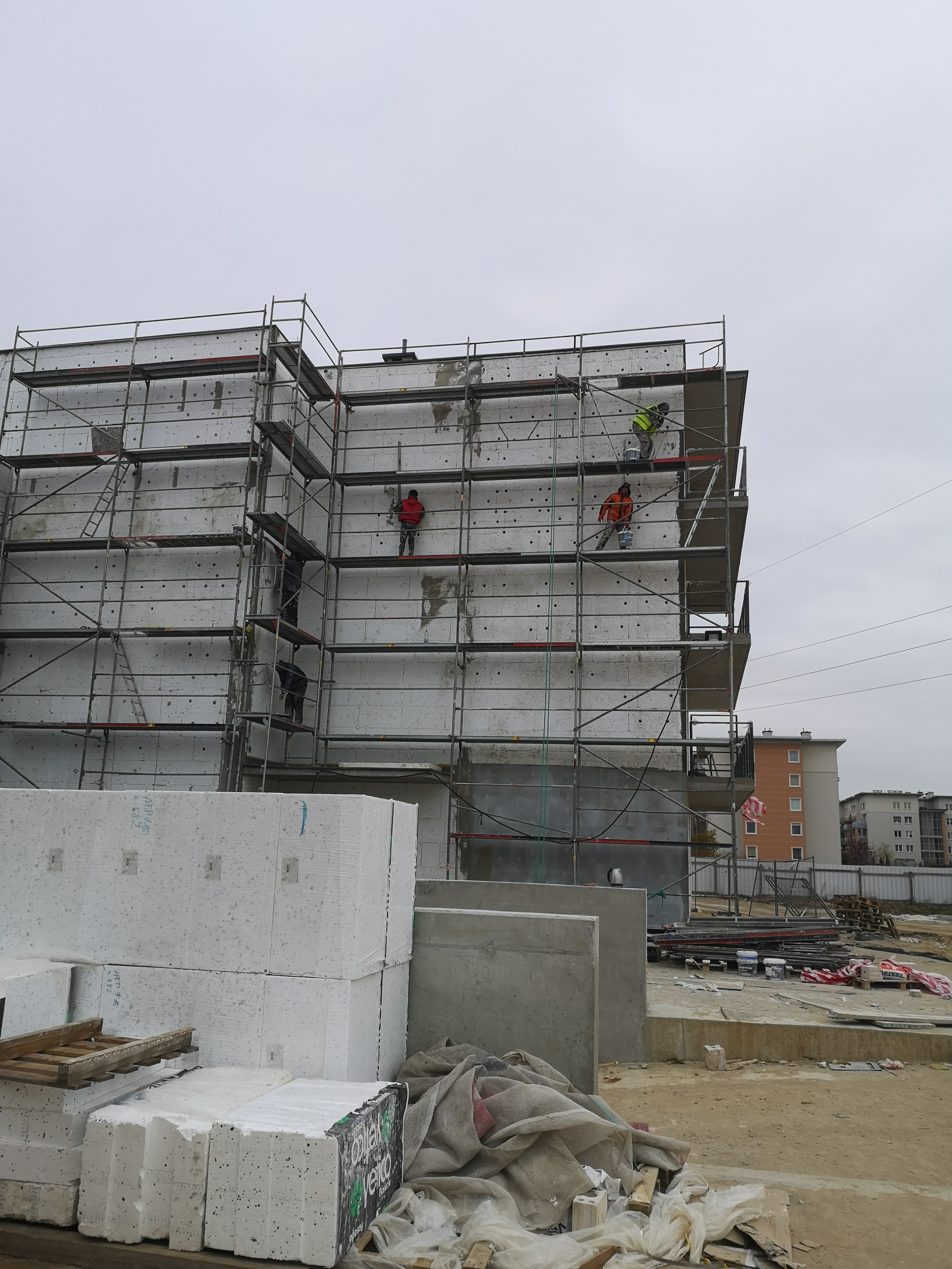 Pracownicy na rusztowaniu podczas ocieplania budynku styropianem, widoczne bloki styropianu na pierwszym planie, pochmurne niebo w tle.