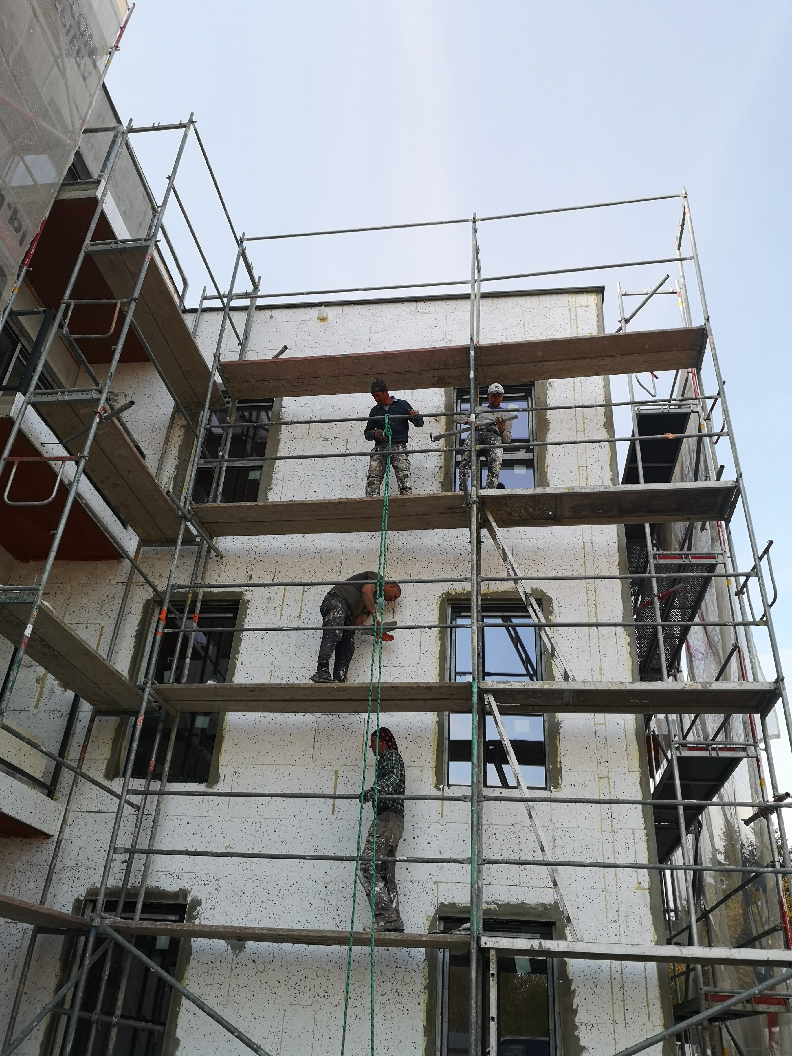 Czterech pracowników na rusztowaniu podczas ocieplania elewacji budynku wielorodzinnego, widok z dołu na fasadę pokrytą styropianem, przygotowywaną do nałożenia tynku.