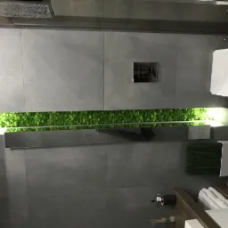 Nowoczesna łazienka z szarymi płytkami, podświetlanym pasem zielonego mchu, minimalistyczną toaletą i drewnianą szafką pod umywalką z białymi ręcznikami.