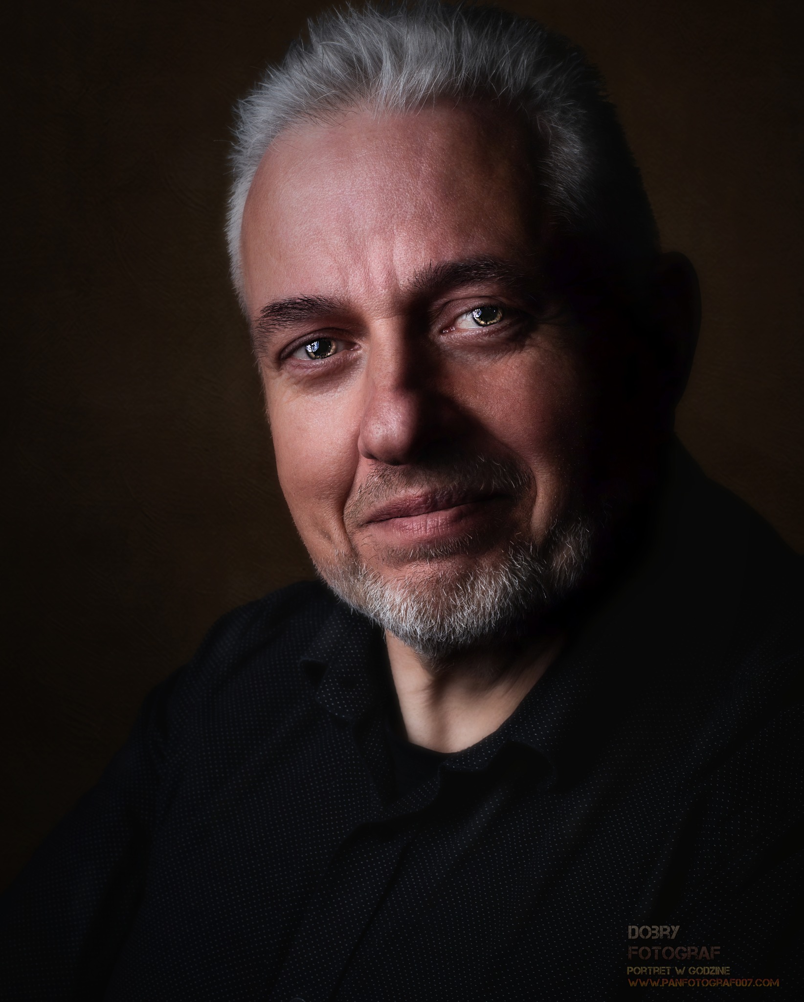 Portret mężczyzny z siwą brodą i włosami, ubranego w ciemną koszulę, wykonany w studiu z ciemnym tłem i delikatnym oświetleniem.