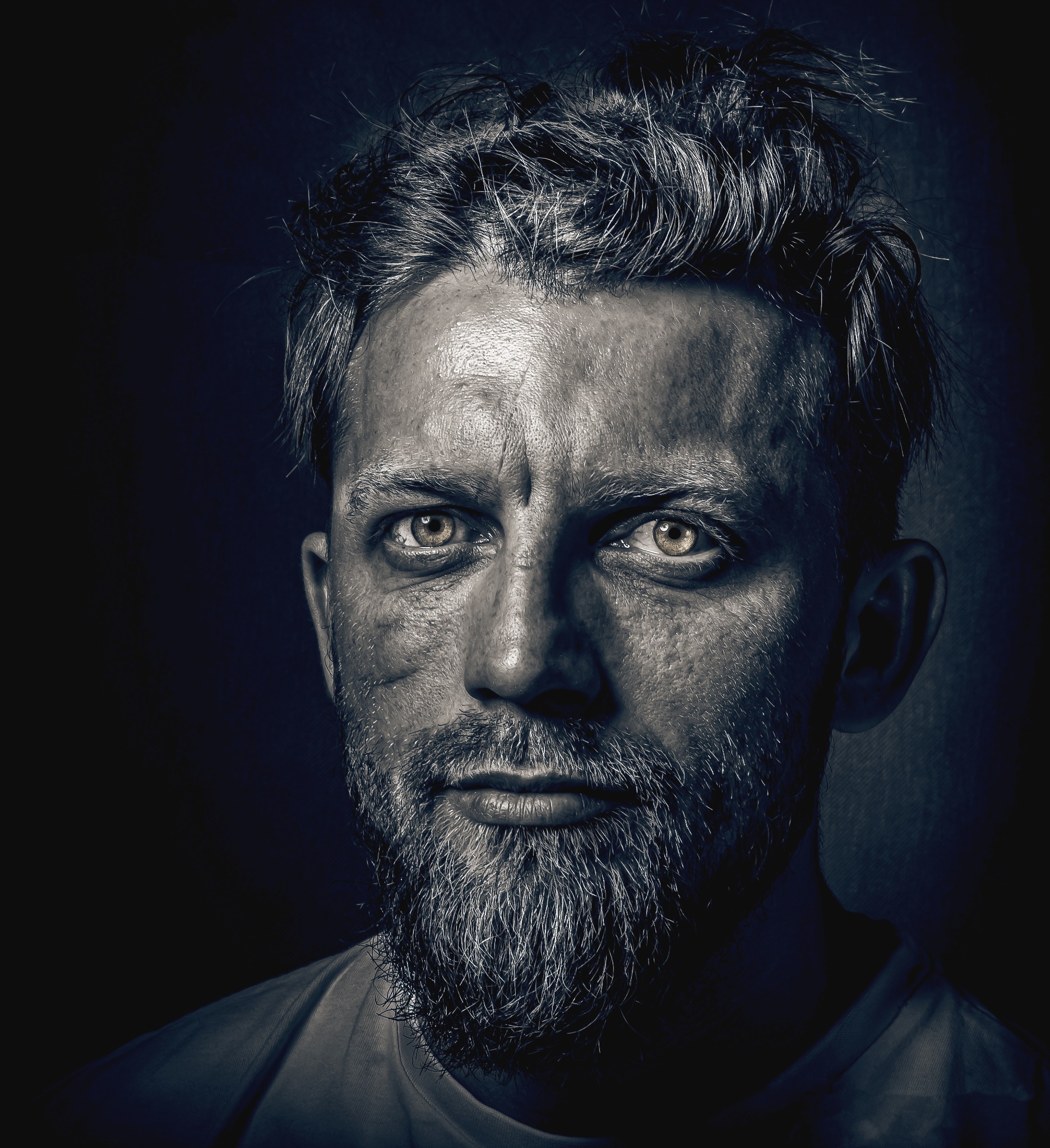 Portret mężczyzny z brodą, oświetlonego z góry, z widocznymi zmarszczkami i blizną na policzku, w ciemnej tonacji.