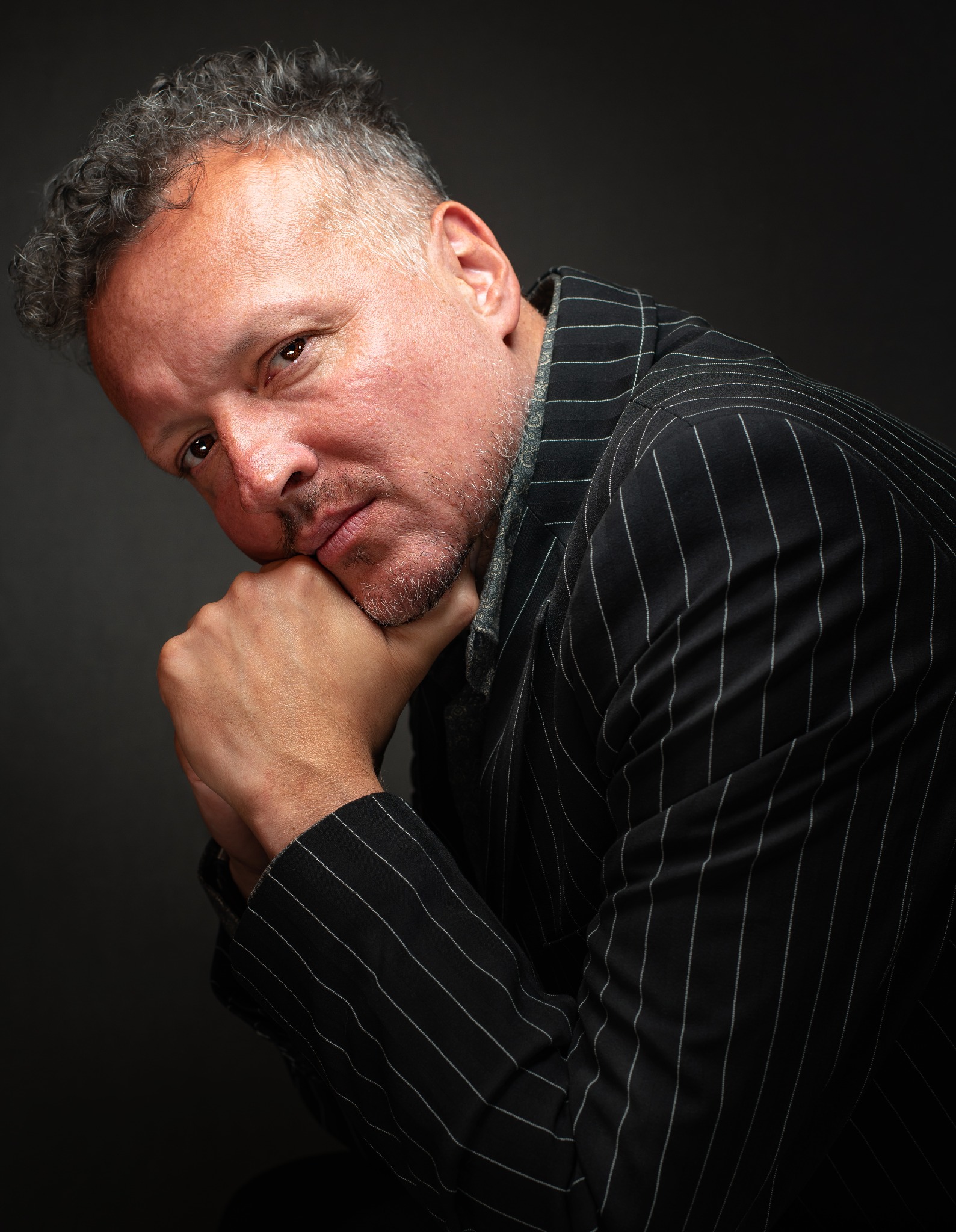 Portret mężczyzny w czarnym garniturze w białe paski, opierającego brodę na dłoniach, z widoczną fakturą skóry i siwiejącymi włosami.