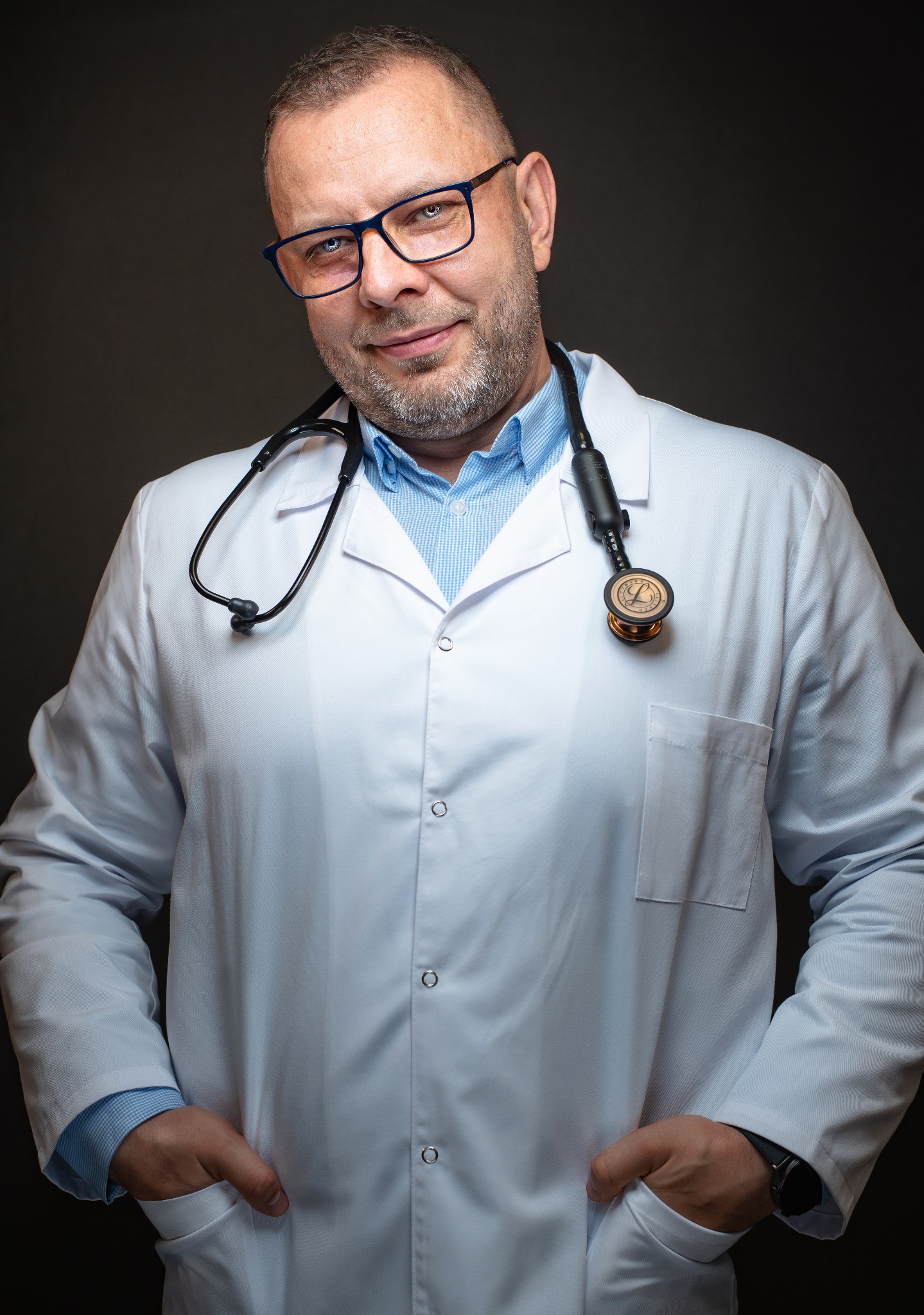 Portret uśmiechniętego lekarza w okularach i białym fartuchu medycznym ze stetoskopem na szyi, z rękami w kieszeniach na tle ciemnego tła.