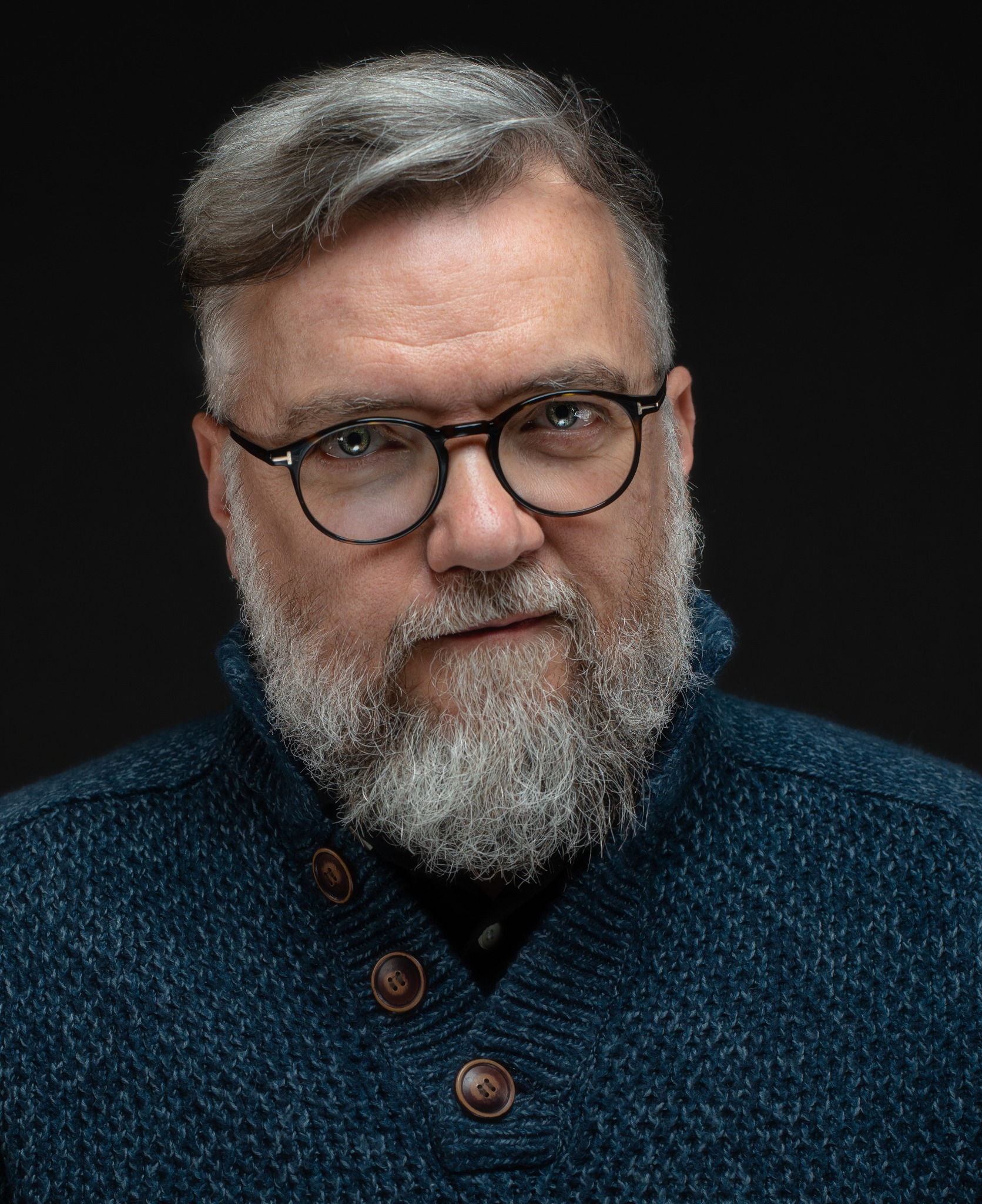 Portret mężczyzny z siwą brodą i włosami, w okularach o okrągłych oprawkach i granatowym swetrze z drewnianymi guzikami, na czarnym tle.