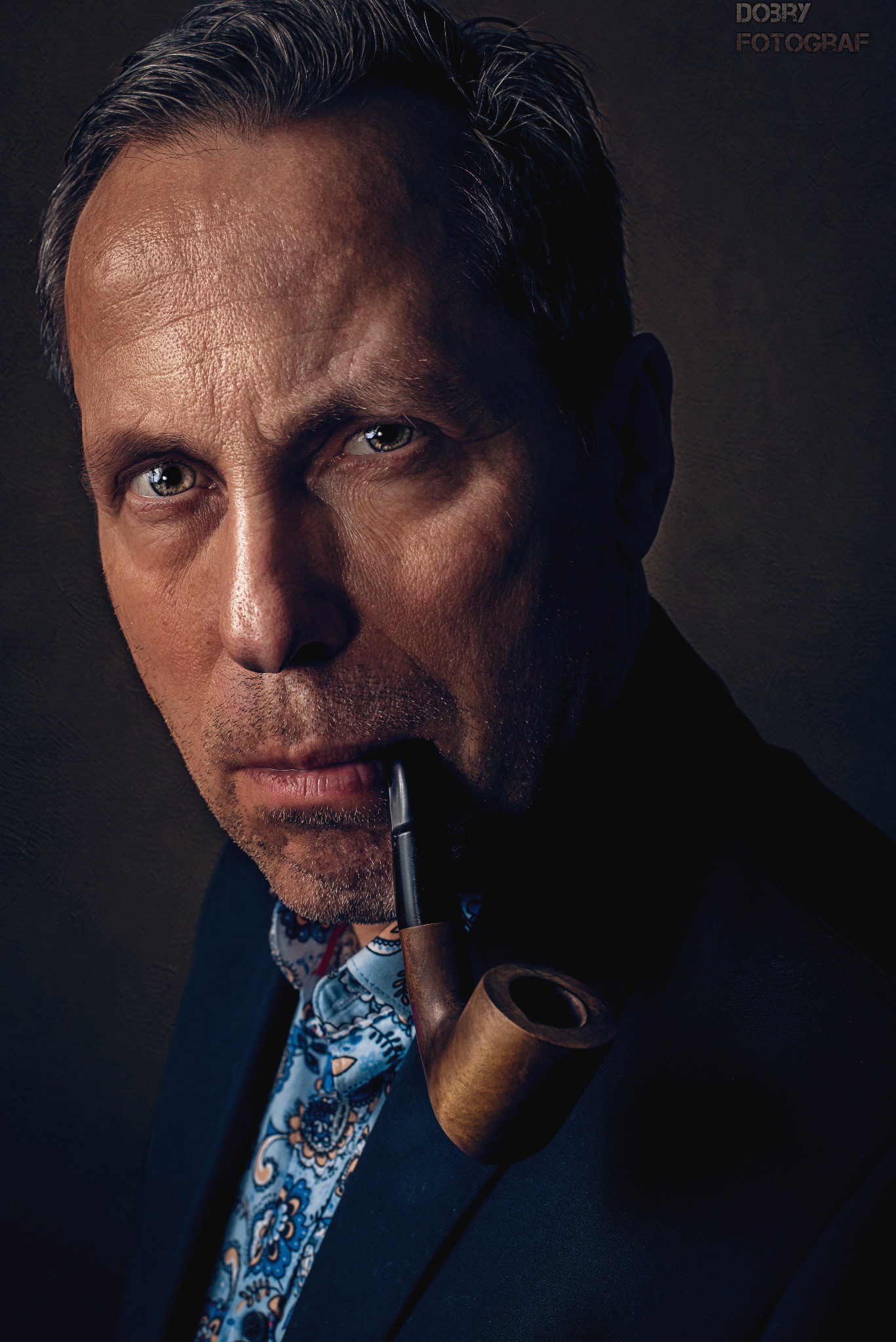 Portret mężczyzny z fajką w ustach, w ciemnym garniturze i wzorzystej koszuli, ostre światło podkreśla rysy twarzy.