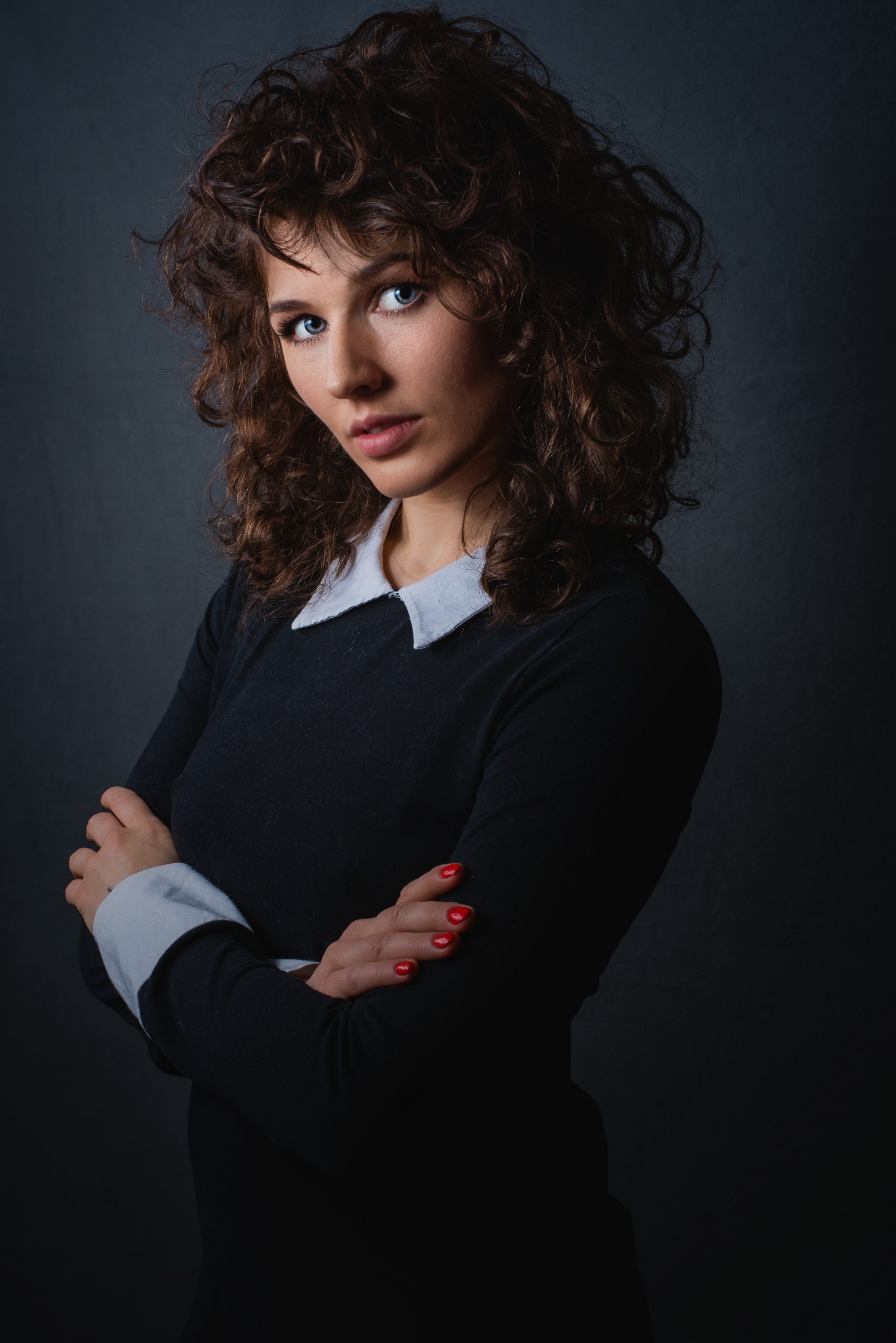 Portret kobiety o kręconych włosach i niebieskich oczach, ubrana w czarną sukienkę z białym kołnierzykiem, pozująca na ciemnym tle z założonymi rękami, z widocznym czerwonym manicure.